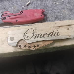 May include: A folding pocket knife with a light-colored wooden handle featuring the engraved word Omertà in a cursive script. The blade is partially visible and displays a row of five cut-out stars. A red utility knife is positioned above it.