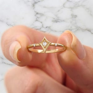May include: A gold ring featuring a central diamond set within a diamond-shaped frame, flanked by two smaller diamonds on each side of the band. The metal has a polished finish, and the design incorporates a geometric, pointed setting style.