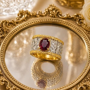 Può includere: Un anello a fascia larga con una gemma rosso scuro dal taglio ovale al centro. L'anello presenta un bordo dorato testurizzato sui bordi superiore e inferiore, con un motivo in filigrana color argento al centro attorno alla pietra.
