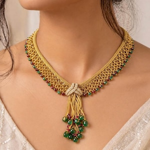 May include: A gold-toned mesh necklace featuring a central knot detail encrusted with clear stones. The collar is accented with small red and green beads along the edges, and a dangling tassel of gold chains tipped with green and red beads.