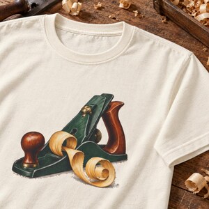 Peut inclure: Un t-shirt à manches courtes de couleur crème arborant une illustration graphique d'un rabot à main vert foncé avec une poignée et un bouton en bois brun poli, accompagné d'un copeau de bois enroulé reposant sur la base de l'outil.