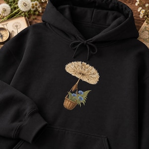 May include: A black hooded sweatshirt featuring a graphic print of a woven basket acting as a hot air balloon, suspended by a large, light brown dandelion seed head. The basket contains green fern leaves and small blue flowers.