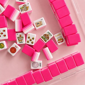 May include: A collection of mahjong tiles featuring bright pink backs and white faces with various colorful illustrations including food items, text labels like NORTH, WEST, JOKER, FLOWER, RED, and SOAP, and numerical symbols, arranged on a pink surface.