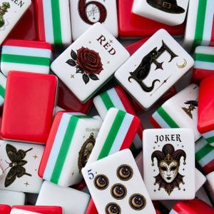 May include: A collection of rectangular mahjong tiles featuring white faces with various illustrations including a red rose, a black cat, a joker mask, a moth, and moon symbols. Some tiles display the text RED, FLOWER, and JOKER with red and green sides.