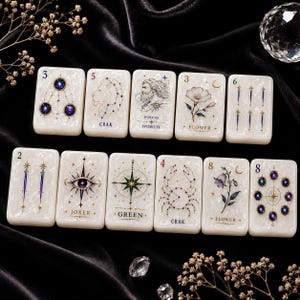 Può includere: Un set di undici tessere rettangolari bianco perlaceo con intricate illustrazioni celesti, floreali e mitologiche in blu, oro e nero. Ogni tessera mostra simboli, numeri e testi unici, tra cui BOREAS NORTH, CRAK, FLOWER, JOKER e GREEN.