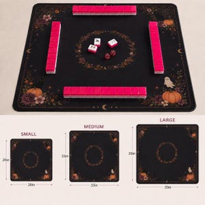 May include: A black square mahjong mat featuring a floral and pumpkin border with a ghost and cat illustration. The mat includes four rows of pink tiles, three loose tiles, and two dice. Three sizes are shown with dimensions of 26, 33, and 39 inches.