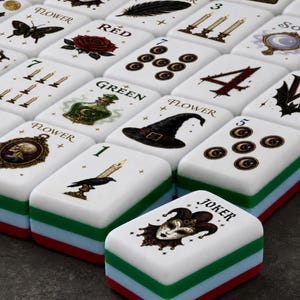May include: A set of rectangular mahjong tiles with white faces featuring various gothic and mystical illustrations including a joker, a rose, a skull, a witch hat, candles, and a raven. Each tile has a layered base of white, green, blue, and red.