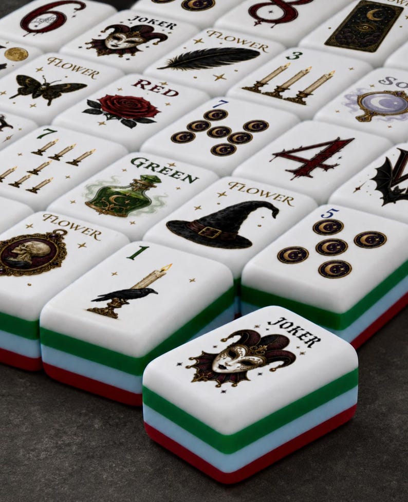 May include: A set of rectangular mahjong tiles with white faces featuring various gothic and mystical illustrations including a joker, a rose, a skull, a witch hat, candles, and a raven. Each tile has a layered base of white, green, blue, and red.