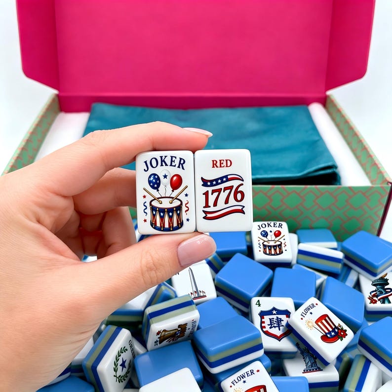 May include: A set of white rectangular Mahjong tiles featuring patriotic American-themed graphics, including a drum with balloons labeled JOKER, the text RED 1776, and various icons like the Statue of Liberty, all housed in a pink and green box.