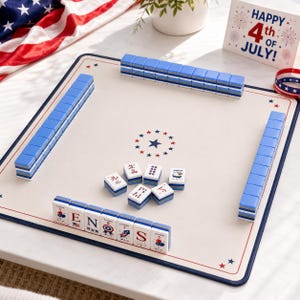 Puede incluir: Un juego de Mahjong con un tapete de color crema que presenta una estrella central y detalles en rojo, blanco y azul. Las fichas est&aacute;n dispuestas en filas con el reverso azul, mientras que varias fichas muestran gr&aacute;ficos patri&oacute;ticos.