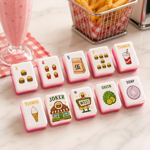 May include: A set of ten white mahjong tiles with pink bases, each featuring colorful food-themed illustrations and text including numbers, Flower, Joker, West, Green, and Soap, arranged on a surface near a milkshake and a basket of fries.