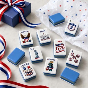 May include: A set of rectangular mahjong tiles featuring patriotic American-themed illustrations, including the Statue of Liberty, the White House, an eagle, and the text 1776, alongside blue gift boxes and red, white, and blue striped ribbons.
