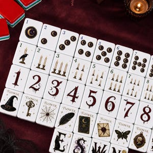 May include: A set of white rectangular mahjong tiles featuring dark, gothic-themed illustrations including crescent moons, lit candles, spiderwebs, keys, witch hats, feathers, moths, and skulls, with numbers and letters printed in a stylized red or black font.
