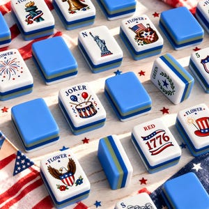 May include: A collection of rectangular mahjong tiles featuring patriotic American-themed illustrations, including the Statue of Liberty, a bald eagle, fireworks, and the year 1776. The tiles have white faces with colorful graphics and blue sides.