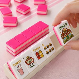 May include: A set of rectangular mahjong tiles with bright pink backs and white faces featuring colorful food-themed illustrations and text including Flower, East, and Joker. The tiles are arranged in a beige rack with several scattered.