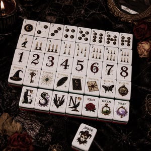 May include: A set of white rectangular mahjong tiles featuring dark gothic illustrations including moons, candles, numbers, flowers, and symbols. The tiles display text such as Flower, E, S, W, N, Red, Gren, Soap, and Joker, arranged in a grid pattern.