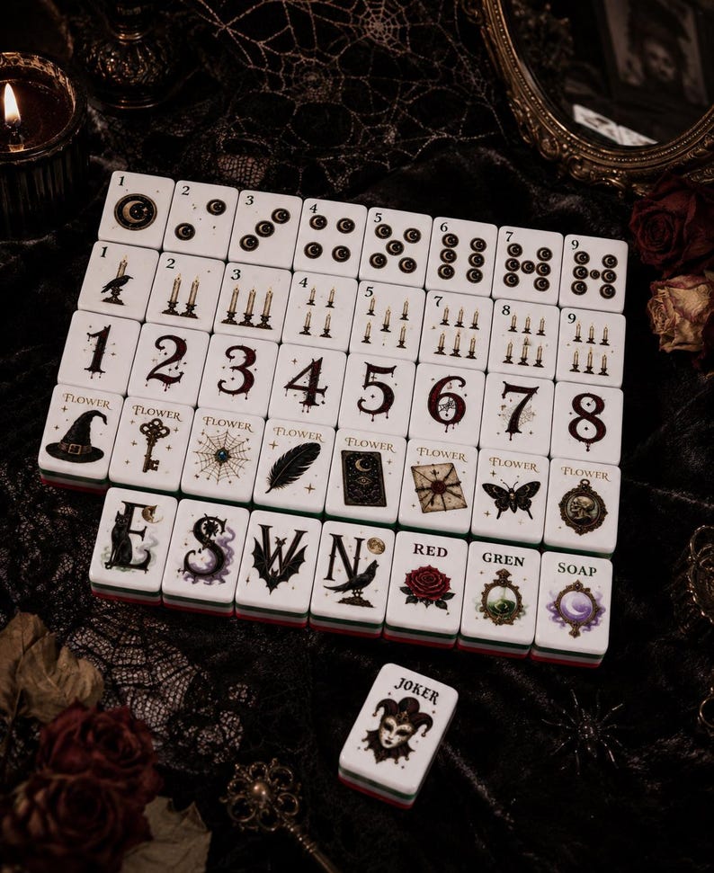 May include: A set of white rectangular mahjong tiles featuring dark gothic illustrations including moons, candles, numbers, flowers, and symbols. The tiles display text such as Flower, E, S, W, N, Red, Gren, Soap, and Joker, arranged in a grid pattern.