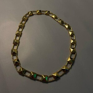 May include: A gold-toned chain necklace featuring a series of repeating pyramid-shaped studs linked together. The metallic links form a circular shape, with each pyramid stud displaying a smooth, reflective surface and a pointed top.