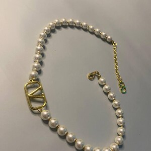 May include: A necklace featuring a strand of white spherical pearls interspersed with small gold-toned beads. A large gold-toned V-shaped logo emblem is integrated into the strand. The necklace includes a gold-toned chain extender with a small V-logo charm.