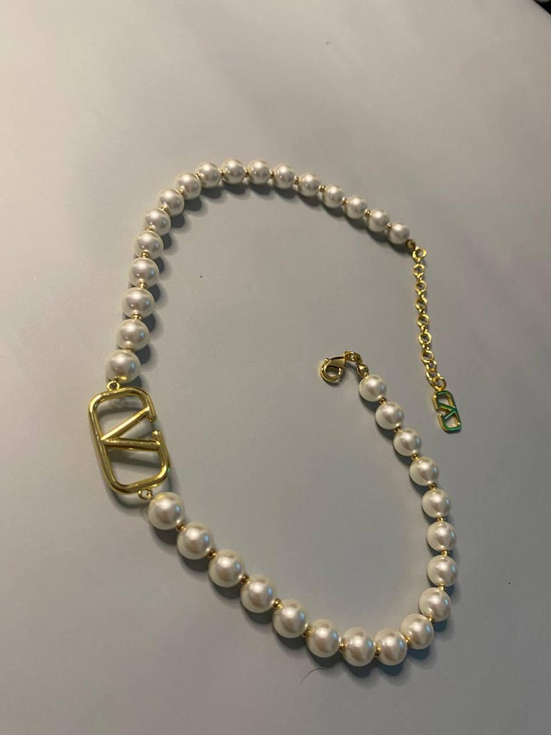 May include: A necklace featuring a strand of white spherical pearls interspersed with small gold-toned beads. A large gold-toned V-shaped logo emblem is integrated into the strand. The necklace includes a gold-toned chain extender with a small V-logo charm.