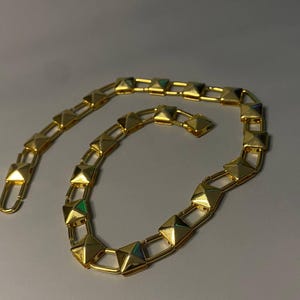 May include: A gold-toned chain necklace featuring a series of repeating square pyramid-shaped studs connected by rectangular metal links. The chain has a polished metallic finish and a hook-style clasp mechanism at one end for secure fastening.