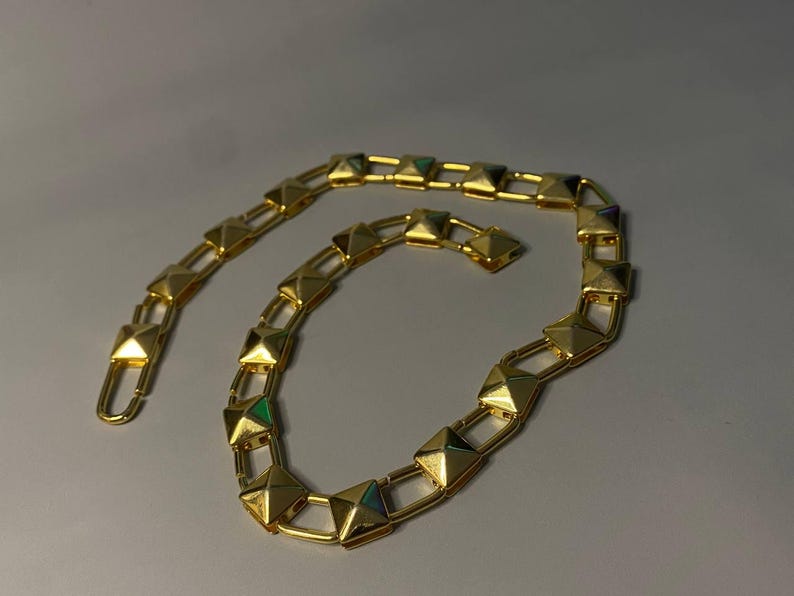 May include: A gold-toned chain necklace featuring a series of repeating square pyramid-shaped studs connected by rectangular metal links. The chain has a polished metallic finish and a hook-style clasp mechanism at one end for secure fastening.
