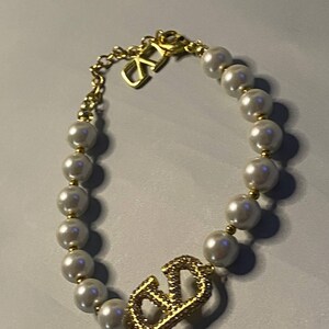 May include: A bracelet featuring a string of white spherical faux pearls interspersed with small gold-toned beads. The piece includes a gold-toned metal charm shaped like the letters V and D, encrusted with clear crystals, and a gold-toned chain link.