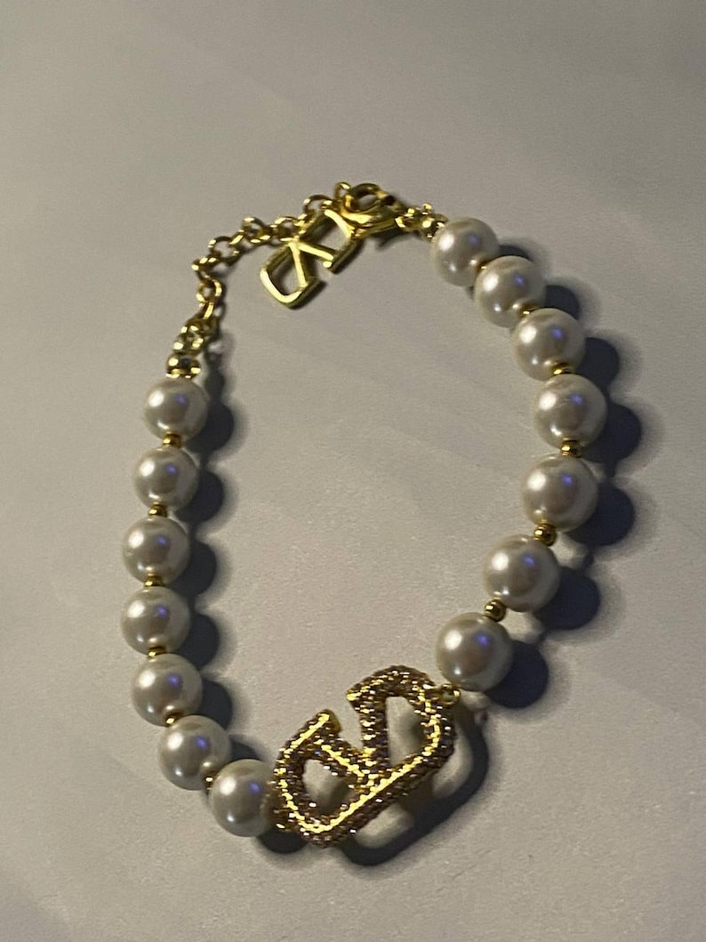 May include: A bracelet featuring a string of white spherical faux pearls interspersed with small gold-toned beads. The piece includes a gold-toned metal charm shaped like the letters V and D, encrusted with clear crystals, and a gold-toned chain link.
