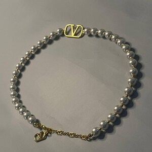 May include: A necklace featuring a strand of white spherical beads interspersed with a gold-toned V-logo emblem. The necklace includes a gold-toned chain segment with a dangling V-logo charm, creating a circular design with metallic accents.