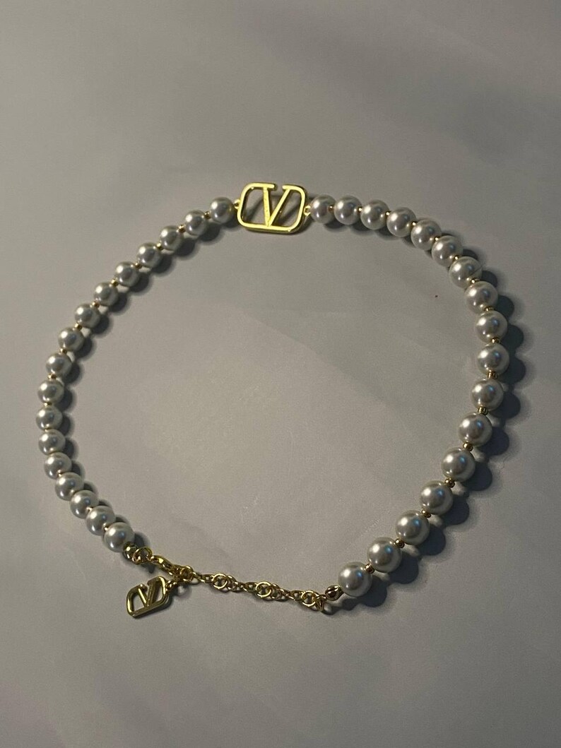 May include: A necklace featuring a strand of white spherical beads interspersed with a gold-toned V-logo emblem. The necklace includes a gold-toned chain segment with a dangling V-logo charm, creating a circular design with metallic accents.