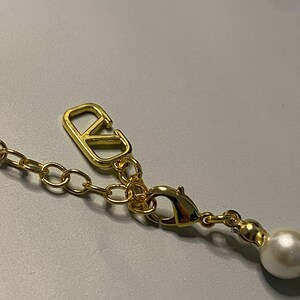May include: A gold-toned metal necklace chain featuring a lobster claw clasp and an extender chain. A polished gold-toned V-shaped logo charm is attached to the chain, and a single round white faux pearl bead hangs from the end of the extender.