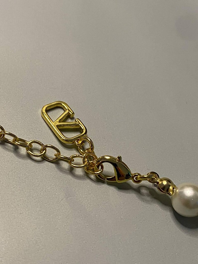 May include: A gold-toned metal necklace chain featuring a lobster claw clasp and an extender chain. A polished gold-toned V-shaped logo charm is attached to the chain, and a single round white faux pearl bead hangs from the end of the extender.
