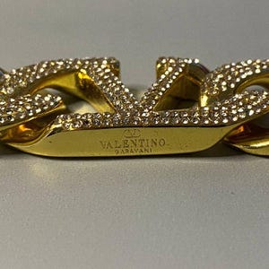 Puede incluir: Una pulsera de eslabones dorados con el logotipo de Valentino Garavani como elemento central. La superficie del logotipo y los eslabones adyacentes est&aacute;n incrustados con peque&ntilde;os cristales claros y reflectantes. El nombre de la marca est&aacute; grabado.