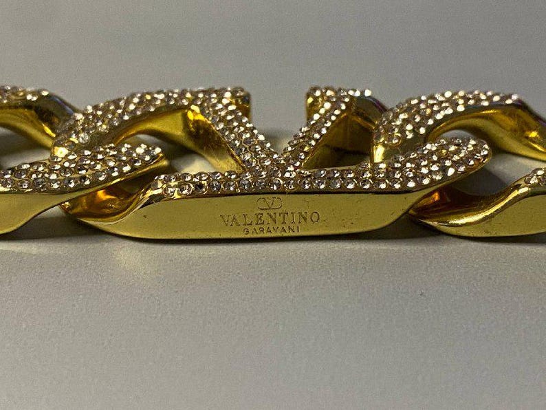 Puede incluir: Una pulsera de eslabones dorados con el logotipo de Valentino Garavani como elemento central. La superficie del logotipo y los eslabones adyacentes est&aacute;n incrustados con peque&ntilde;os cristales claros y reflectantes. El nombre de la marca est&aacute; grabado.