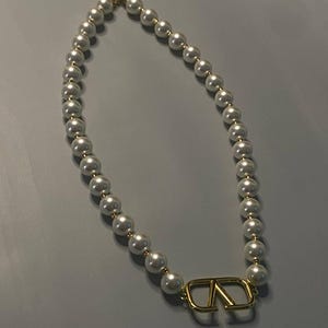 May include: A necklace featuring a strand of white spherical beads interspersed with small gold-toned spacers. The piece includes a prominent gold-toned metal V-shaped logo pendant at the center and a gold-toned chain extension with a clasp.