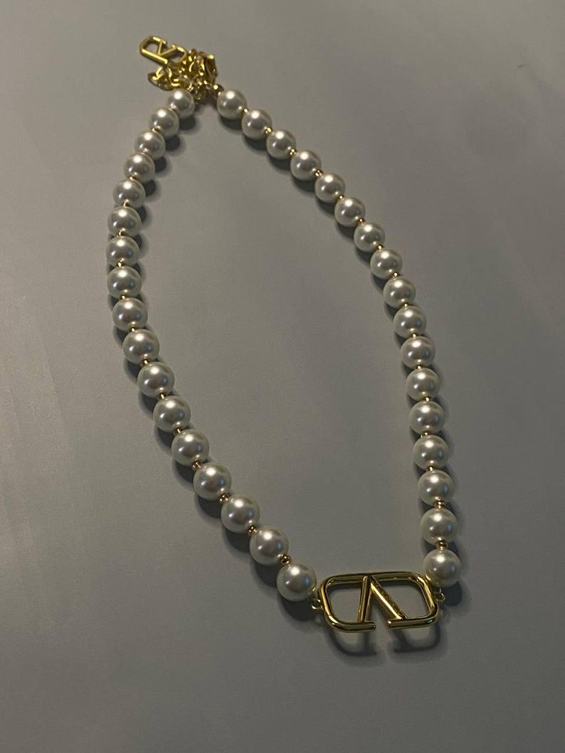May include: A necklace featuring a strand of white spherical beads interspersed with small gold-toned spacers. The piece includes a prominent gold-toned metal V-shaped logo pendant at the center and a gold-toned chain extension with a clasp.