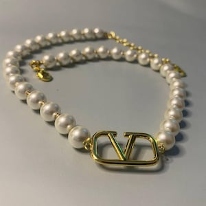 May include: A necklace featuring a strand of white spherical pearls and a central gold-toned metal V-shaped logo charm. The piece includes a gold-toned metal chain extension with a small circular tag at the end, secured by a lobster claw clasp.