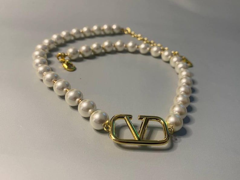 May include: A necklace featuring a strand of white spherical pearls and a central gold-toned metal V-shaped logo charm. The piece includes a gold-toned metal chain extension with a small circular tag at the end, secured by a lobster claw clasp.