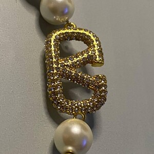 May include: A necklace featuring a series of round white pearls separated by small gold-toned beads, with a prominent gold-toned letter E pendant encrusted with numerous small clear crystals positioned between the pearls.