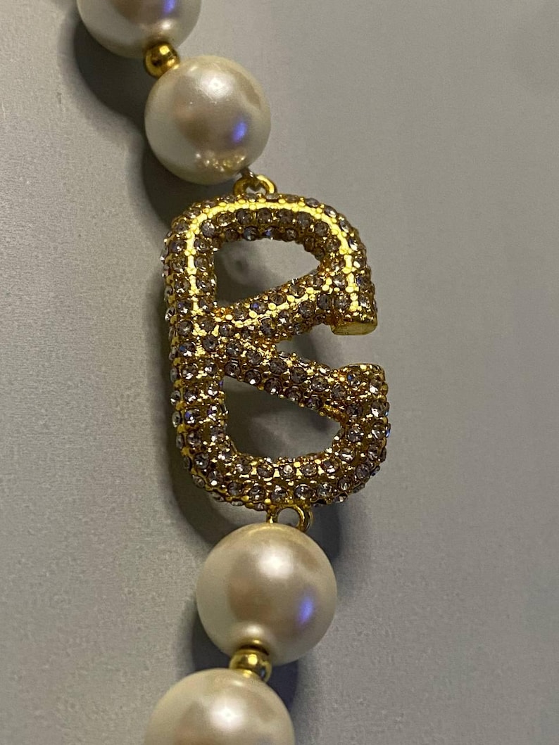 May include: A necklace featuring a series of round white pearls separated by small gold-toned beads, with a prominent gold-toned letter E pendant encrusted with numerous small clear crystals positioned between the pearls.