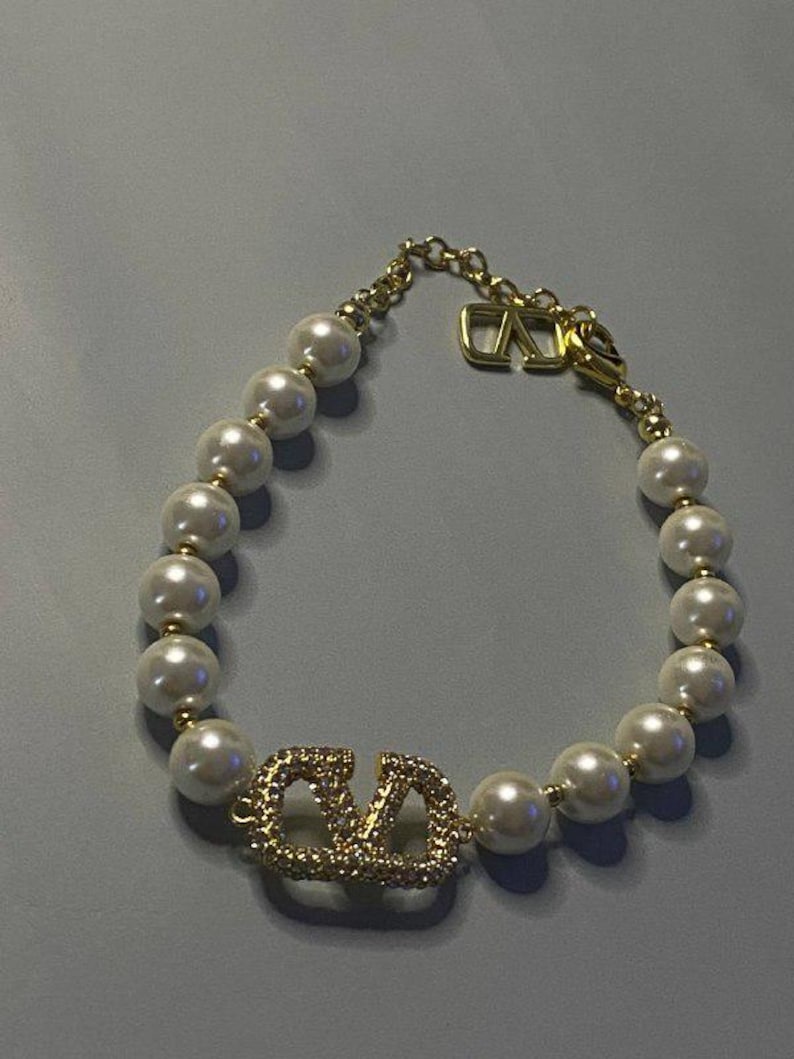 May include: A bracelet featuring a series of white spherical faux pearls separated by small gold-toned beads. The centerpiece is a gold-toned V-shaped logo encrusted with clear crystals. A gold-toned chain with a V-shaped charm serves as the closure.