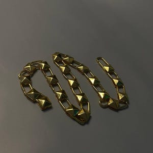 May include: A gold-toned chain necklace featuring a series of repeating pyramid-shaped studs. The chain is constructed from interconnected rectangular links, each holding a raised, four-sided pyramid element with a polished, reflective metallic finish.