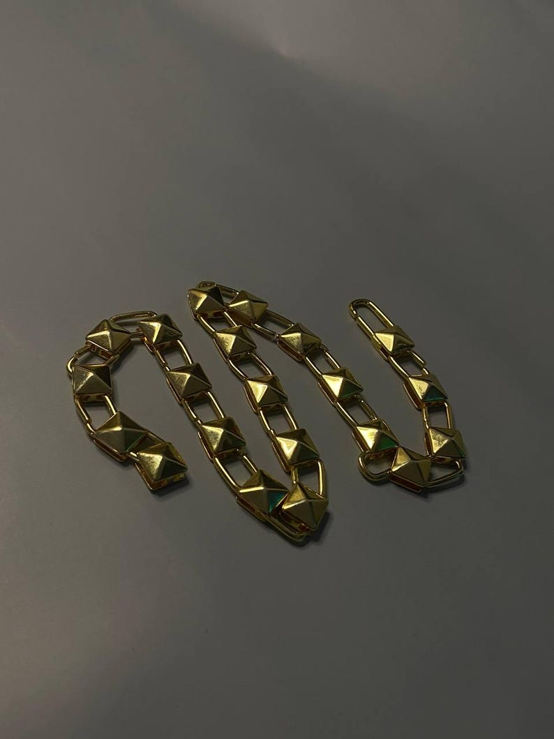 May include: A gold-toned chain necklace featuring a series of repeating pyramid-shaped studs. The chain is constructed from interconnected rectangular links, each holding a raised, four-sided pyramid element with a polished, reflective metallic finish.