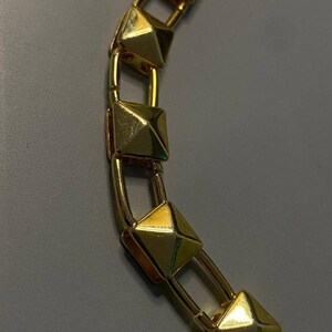May include: A gold-toned chain necklace featuring a series of repeating square pyramid-shaped studs. Each pyramid stud is connected by smooth, rounded gold-coloured metal links, creating a structured and uniform design along the entire length.