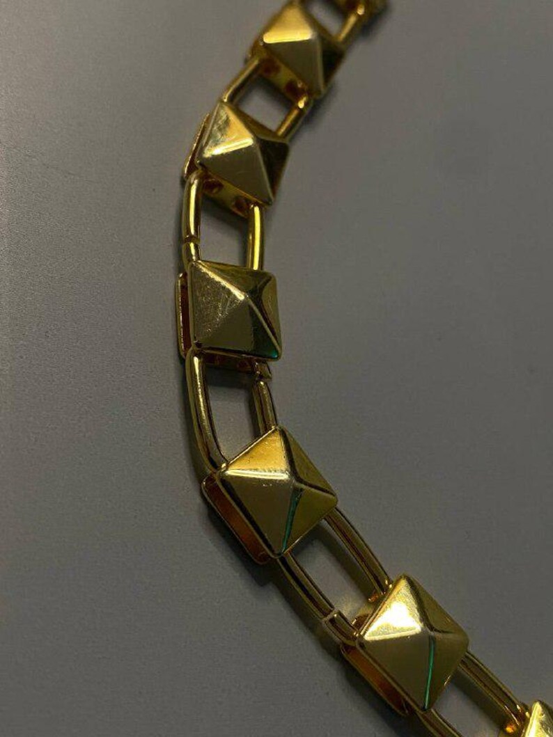 May include: A gold-toned chain necklace featuring a series of repeating square pyramid-shaped studs. Each pyramid stud is connected by smooth, rounded gold-coloured metal links, creating a structured and uniform design along the entire length.