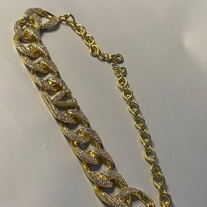Puede incluir: Un collar de cadena dorada que presenta un segmento de cadena gruesa tipo barbada incrustado con brillantes transparentes en los eslabones frontales, conectado a una cadena m&aacute;s fina de eslabones ovalados lisos con cierre de mosquet&oacute;n.