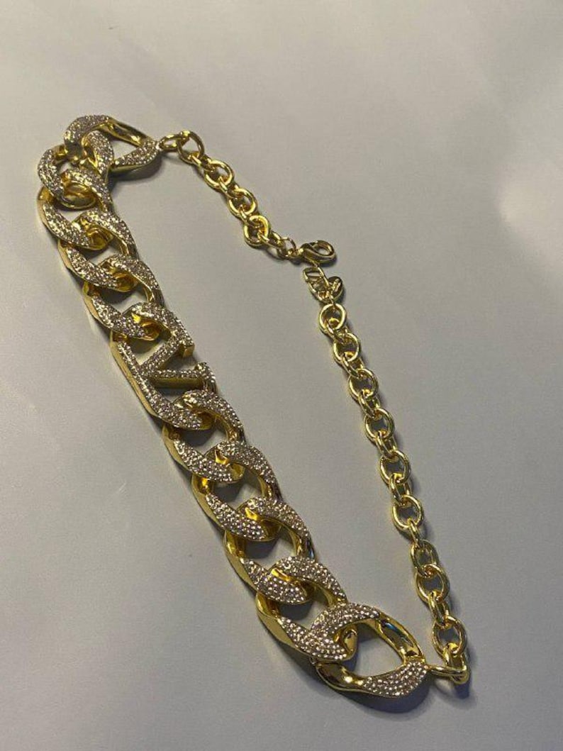Puede incluir: Un collar de cadena dorada que presenta un segmento de cadena gruesa tipo barbada incrustado con brillantes transparentes en los eslabones frontales, conectado a una cadena m&aacute;s fina de eslabones ovalados lisos con cierre de mosquet&oacute;n.