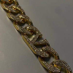 Puede incluir: Un collar de cadena dorada con eslabones gruesos y entrelazados. Cada eslab&oacute;n est&aacute; incrustado con peque&ntilde;as piedras claras y brillantes. Un eslab&oacute;n central prominente muestra las letras VLTN, tambi&eacute;n cubiertas de piedras, con la palabra VALENTINO grabada.