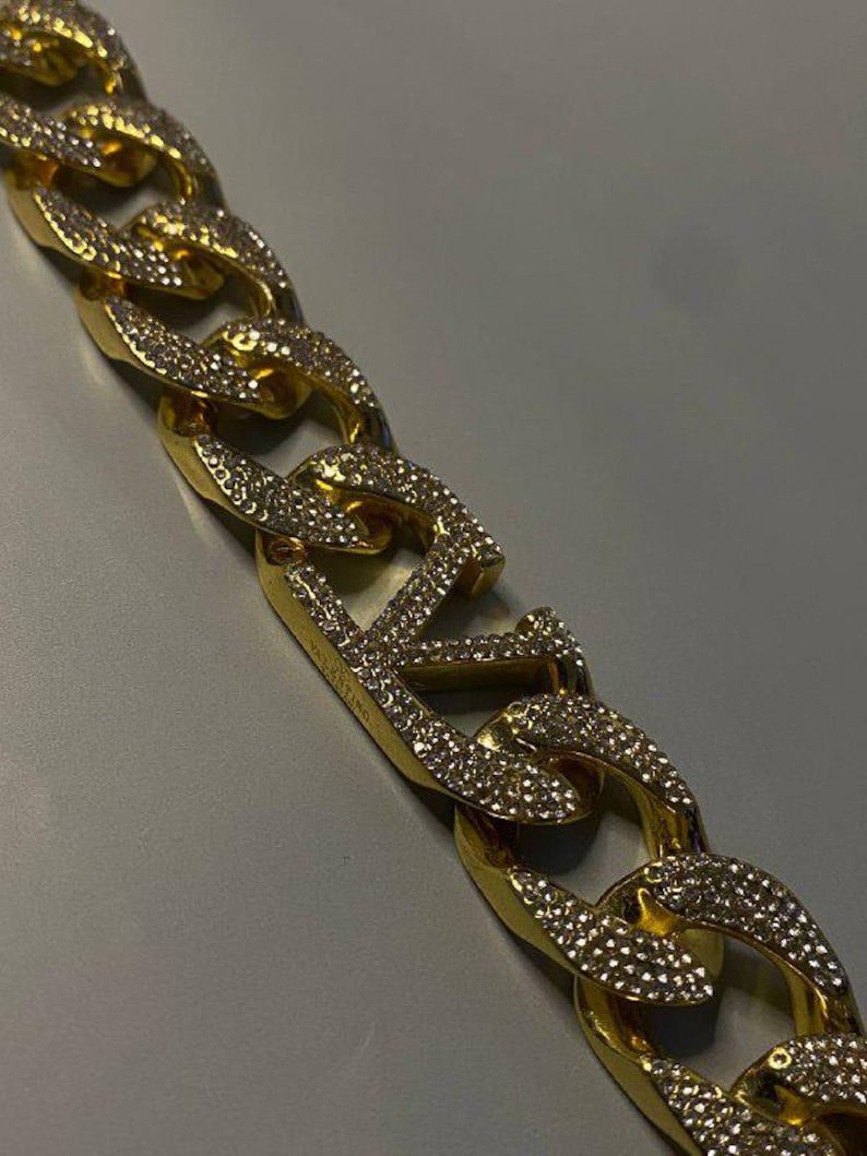 Puede incluir: Un collar de cadena dorada con eslabones gruesos y entrelazados. Cada eslab&oacute;n est&aacute; incrustado con peque&ntilde;as piedras claras y brillantes. Un eslab&oacute;n central prominente muestra las letras VLTN, tambi&eacute;n cubiertas de piedras, con la palabra VALENTINO grabada.