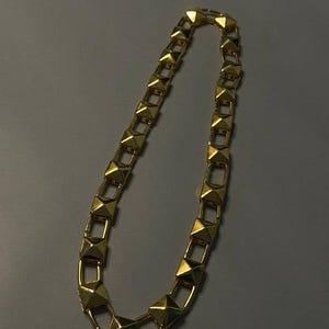 May include: A gold-toned chain necklace featuring a series of repeating pyramid-shaped studs linked together. The metallic links are square-shaped and connected by small rectangular bars, creating a continuous, uniform geometric pattern throughout.